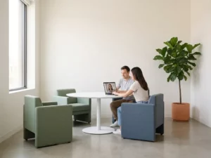 Two colleagues collaborating at a white table in a sunlit office lounge, with a remote teammate visible on a laptop screen nearby.