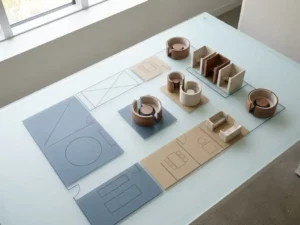 Esquisse minimaliste d'un plan d'étage de bureaux en open space sur du verre dépoli, avec des maquettes architecturales d'espaces collaboratifs et de cabines de concentration dans des tons bleu ardoise et sable chaud.